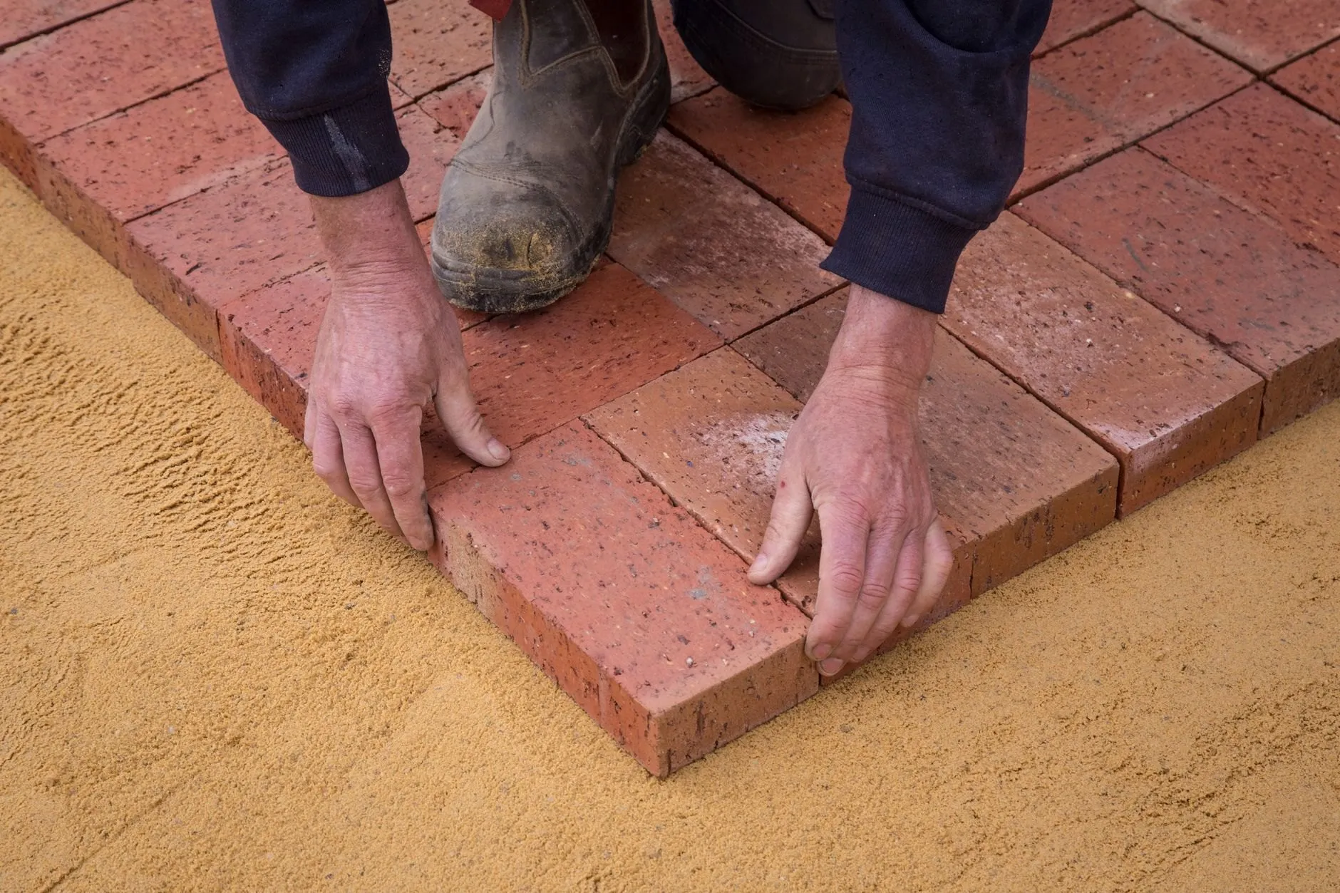 Hands setting reclaimed clay pavers into a sand bed