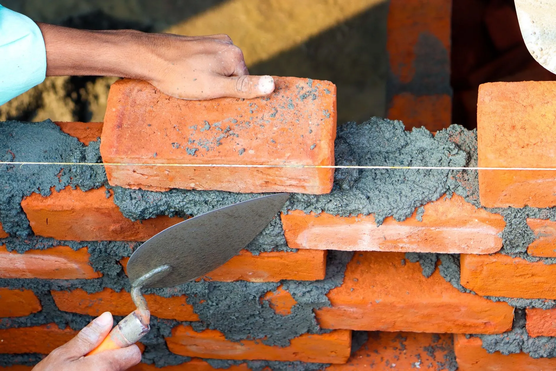 Hands setting a brick onto a fresh mortar bed along a string line, a trowel in the other hand