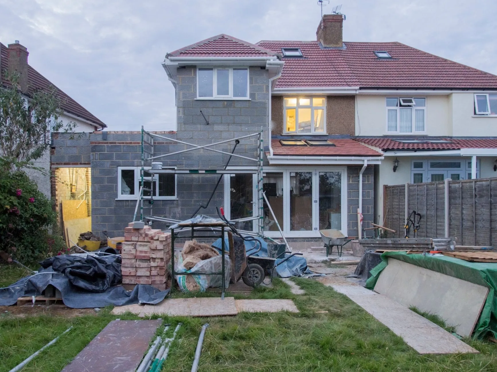 The back of a semi-detached house mid-build: a two-storey side extension in blockwork, scaffold tower, bricks and materials stacked on the lawn