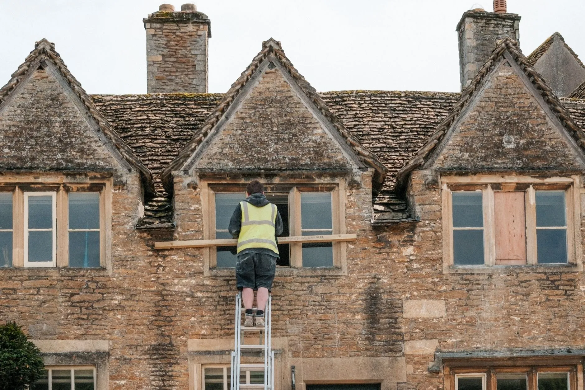 A builder in a hi-vis vest on a ladder repairing the window of a stone cottage with gabled dormers, seen from behind